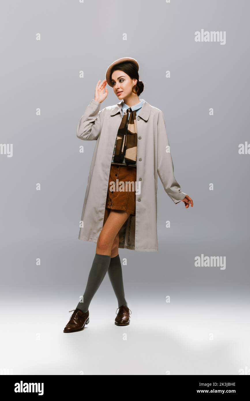 full length of elegant young woman in trench coat adjusting beret on grey,stock image Stock Photo