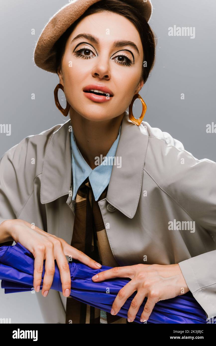 portrait of brunette woman in beret and coat posing with blue umbrella ...