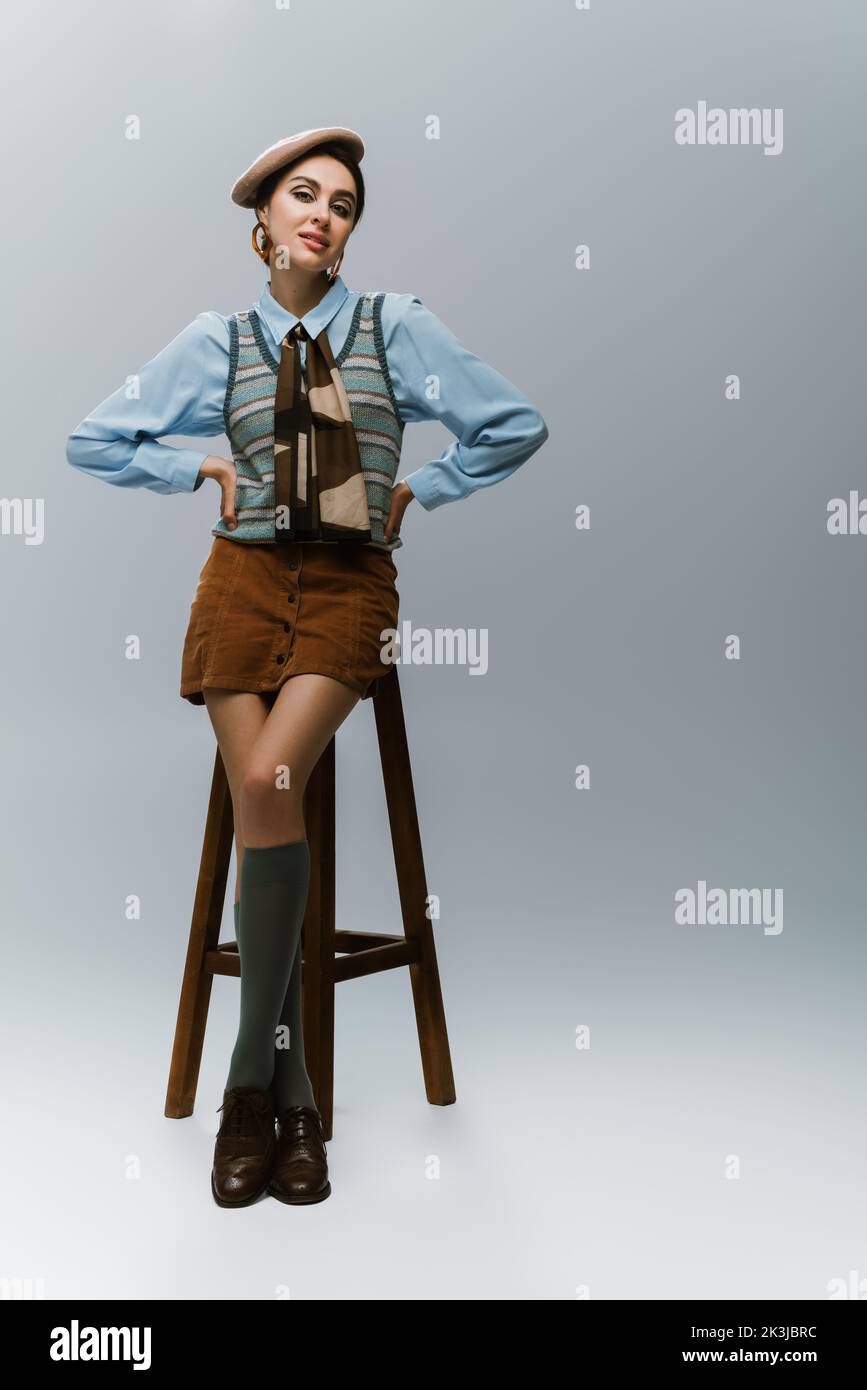 full length of young woman in beret and autumnal clothes standing with hands on hips near wooden high chair on grey,stock image Stock Photo