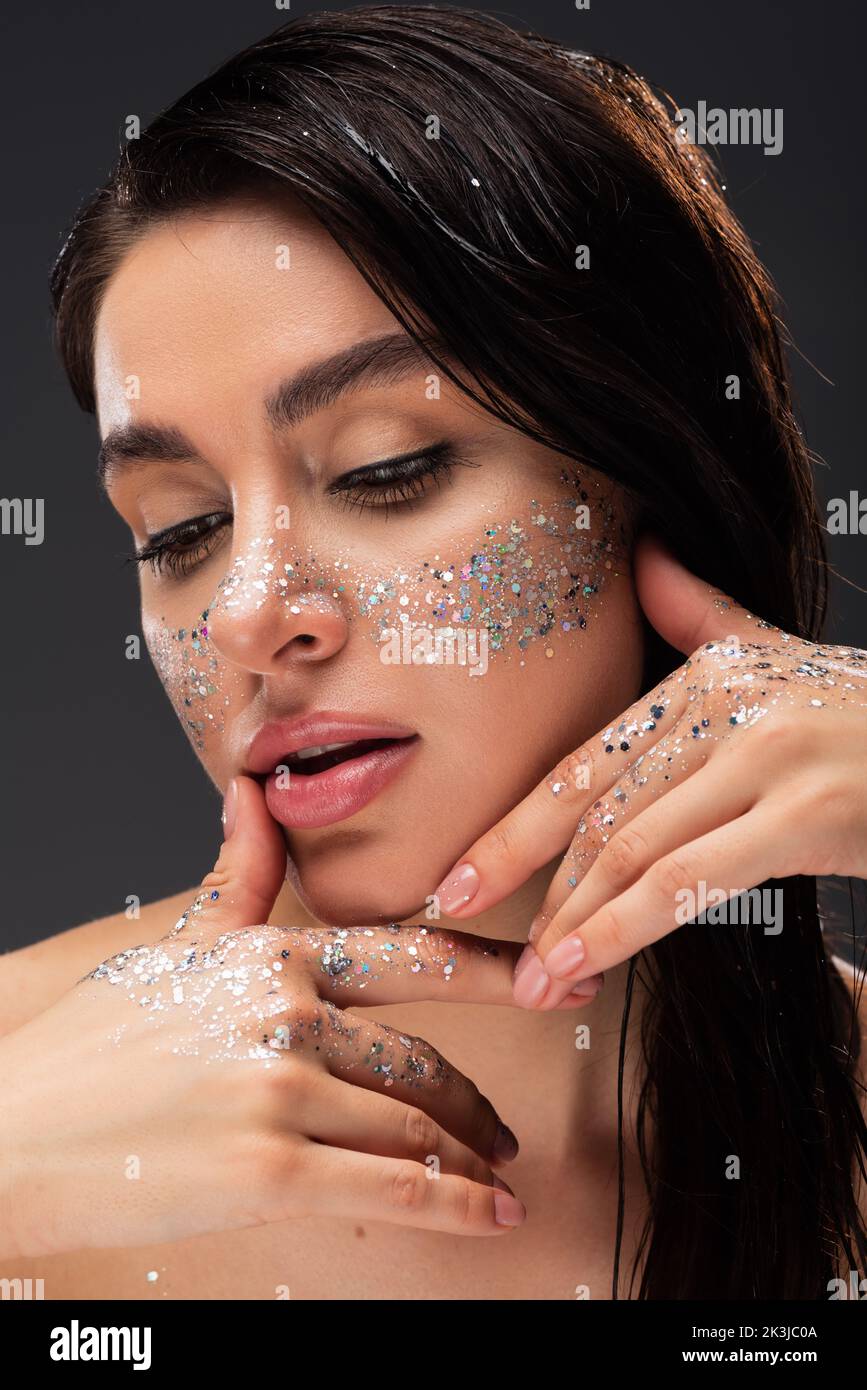 young brunette woman with sparkling glitter on cheeks and hands touching face isolated on grey,stock image Stock Photo