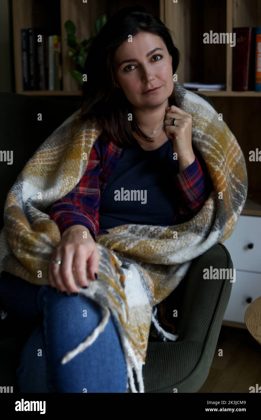 Vertical mature thoughtful, calm brunette woman, covered by blanket, looking at camera and sit in armchair in library Stock Photo