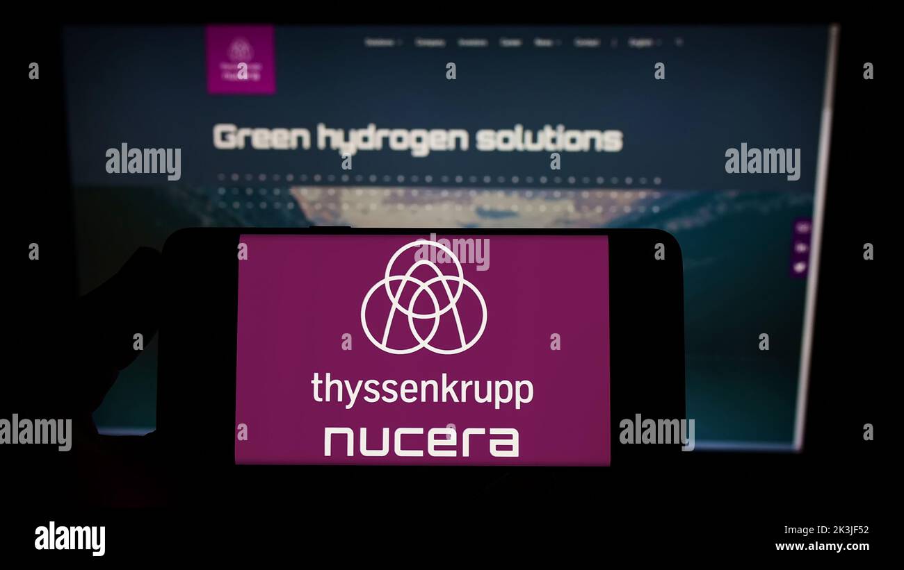 Person holding smartphone with logo of company thyssenkrupp nucera AG Co. KGaA on screen in front of website. Focus on phone display. Stock Photo
