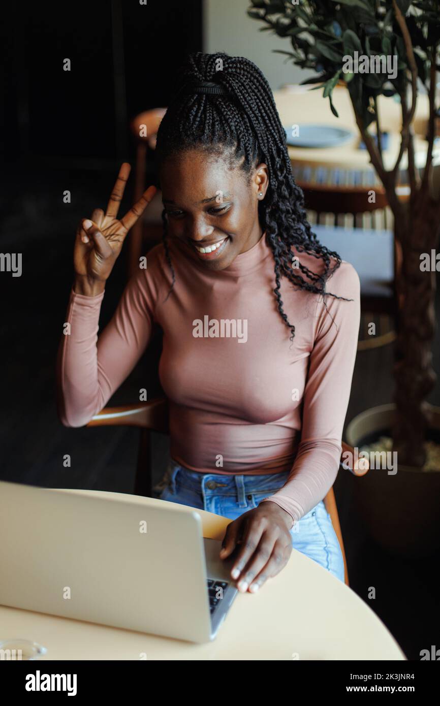 Portrait of young African-American woman sitting at table in cafe near laptop, showing v-sign victory sign with fingers. Stock Photo