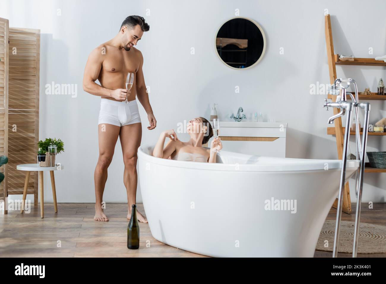 happy woman relaxing in bathtub and looking at boyfriend in underpants standing with champagne glass Stock Photo