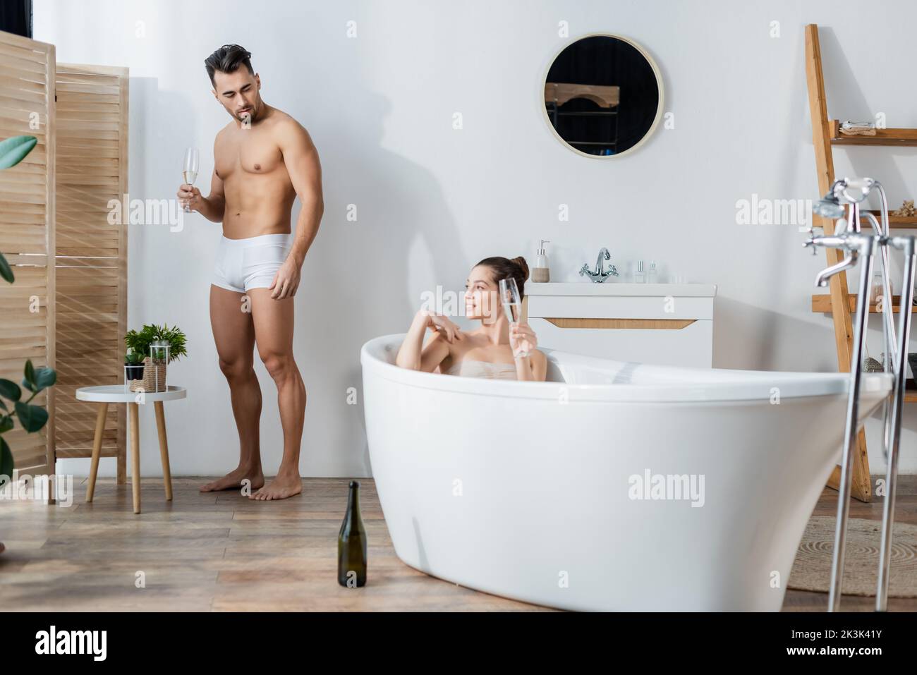 smiling woman relaxing in bathtub near boyfriend in underpants standing with champagne glass Stock Photo