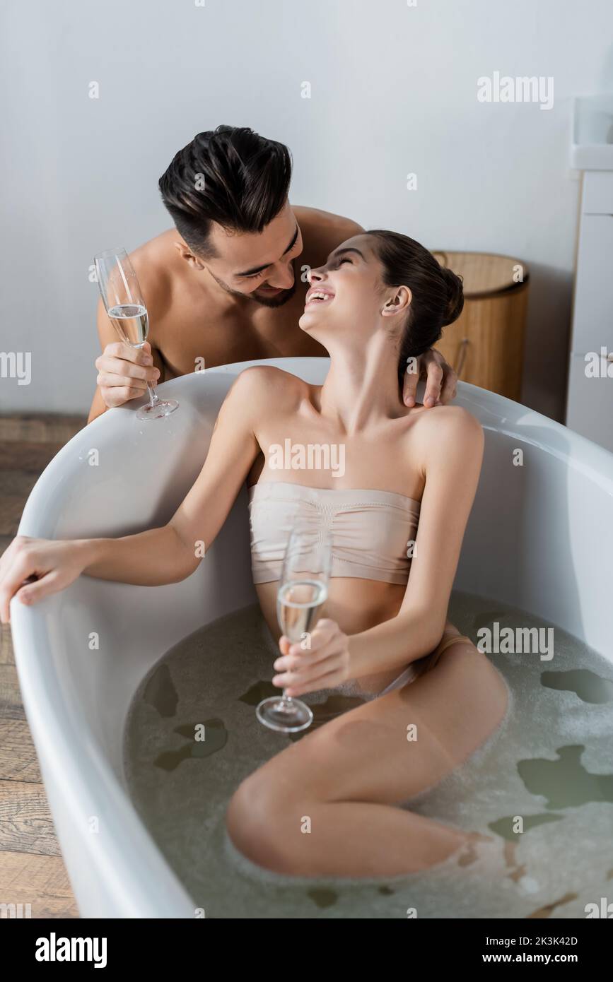 joyful woman taking bath and looking at sexy boyfriend holding champagne glass Stock Photo
