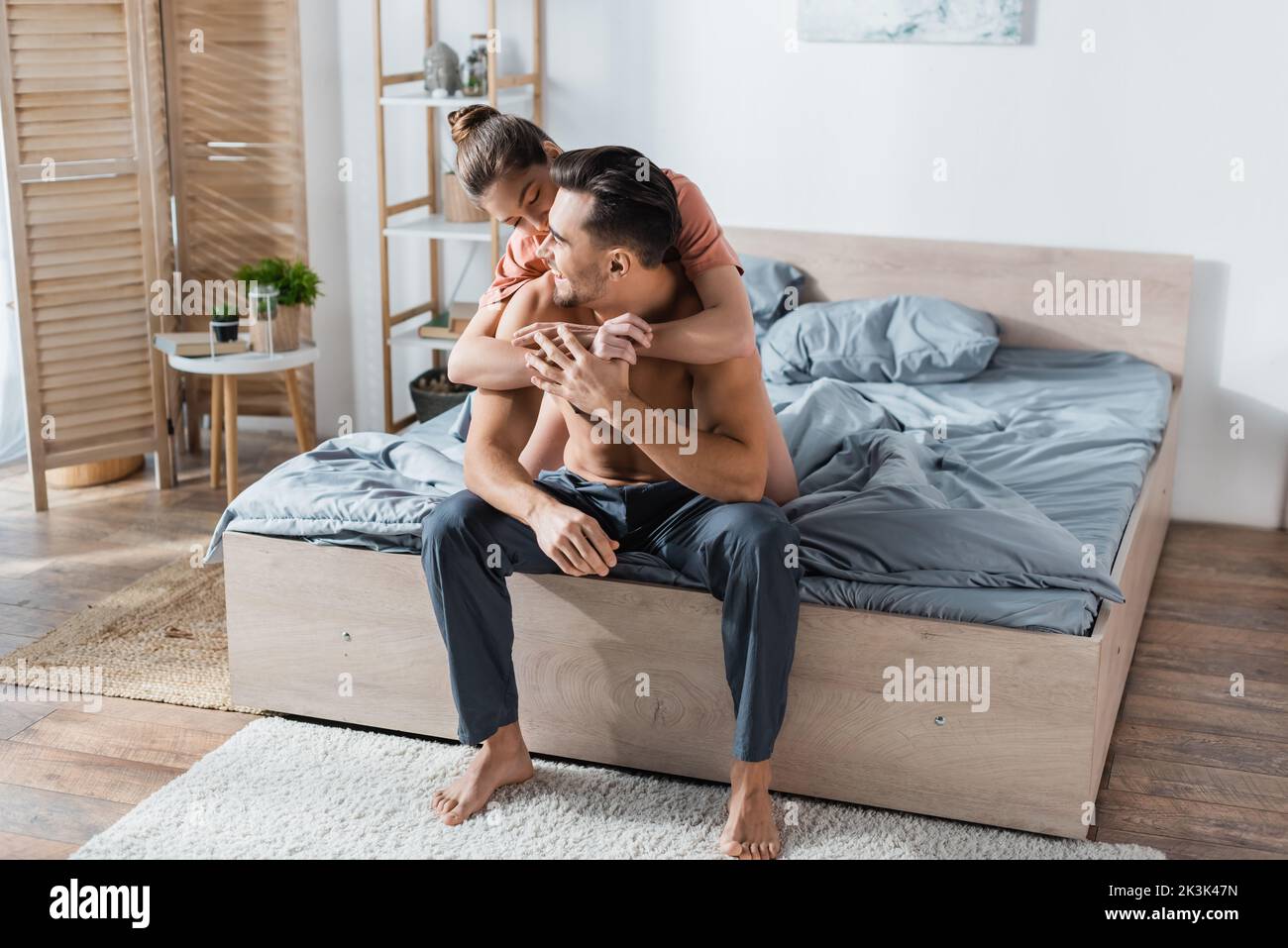 happy shirtless man sitting on bed in pajama pants near girlfriend embracing him in bedroom Stock Photo