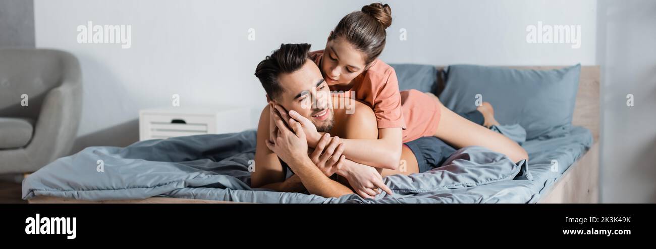 young and hot woman embracing happy boyfriend lying on grey bedding, banner Stock Photo