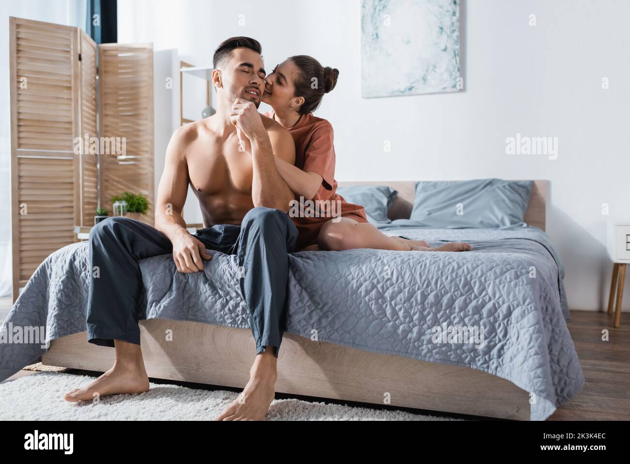 sexy woman in t-shirt kissing shirtless man sitting on bed in pajama pants Stock Photo