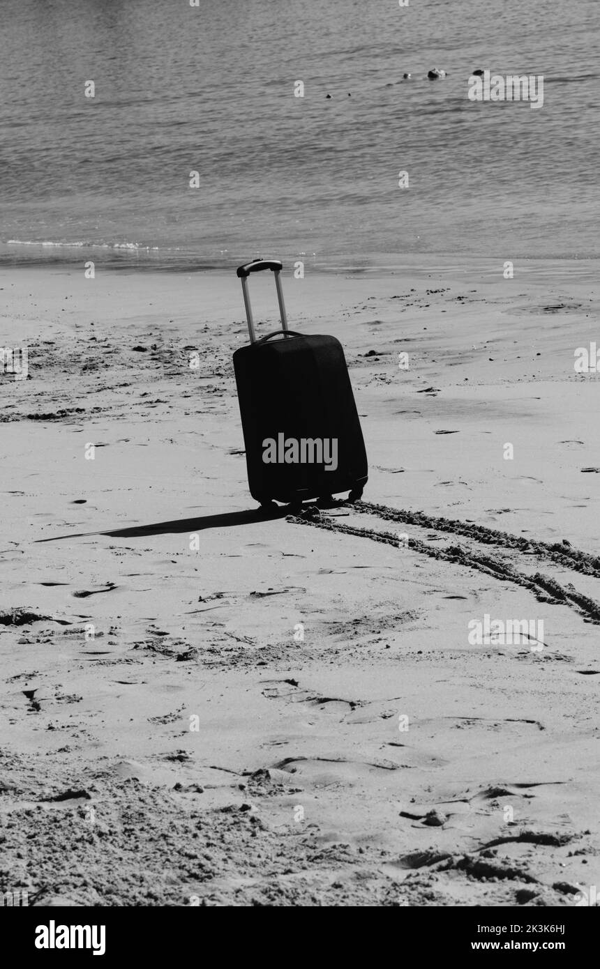 Black travel suitcase on sandy beach with turquoise sea background, summer holidays concept in monochrome Stock Photo