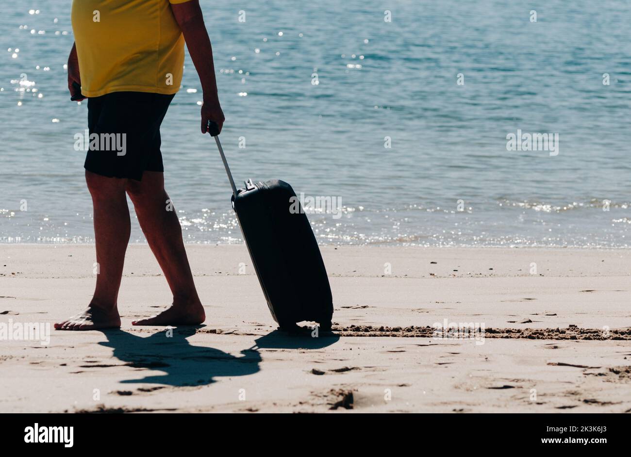 Barefood man wearing shorts and tshirt carrying a black travel suitcase on sandy beach with turquoise sea background, summer holidays concept Stock Photo