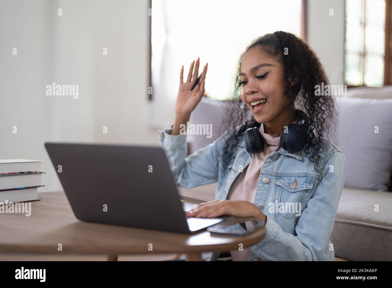 Young woman holds an online meeting while sitting in home. Woman waving hand greeting participants of a video call. Online learning concept. Stock Photo