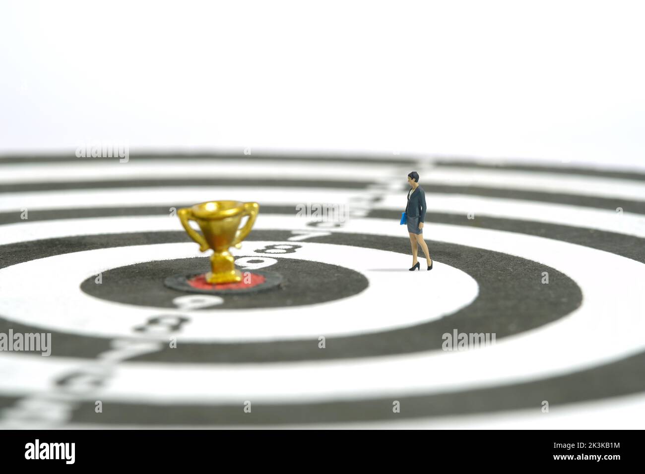 Miniature people toy figure photography. A businesswoman walking above dartboard with gold trophy at the center, isolated on white background. Image p Stock Photo