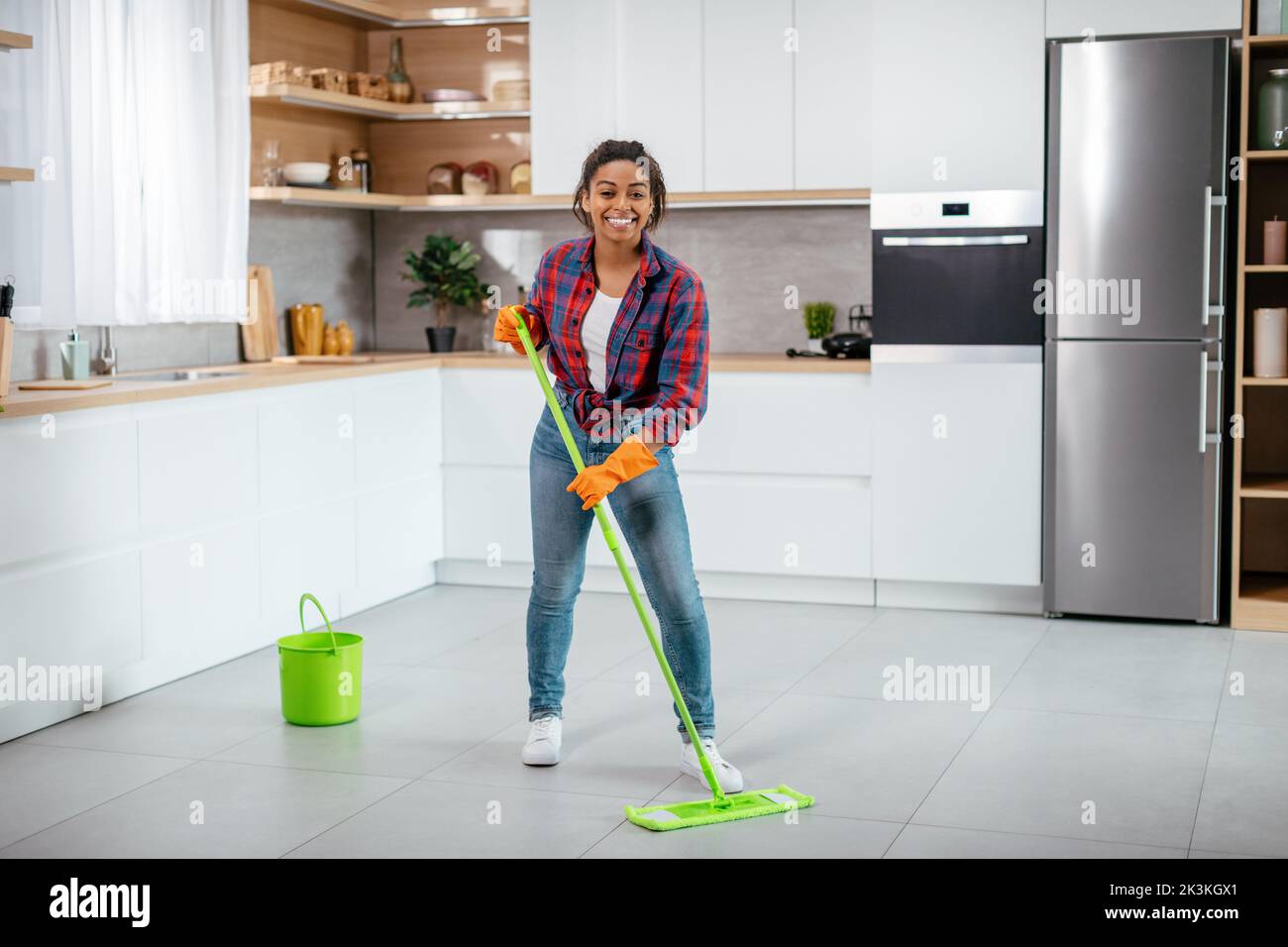 Happy young black woman in rubber gloves washes floor with mop in minimalist kitchen interior, full length Stock Photo