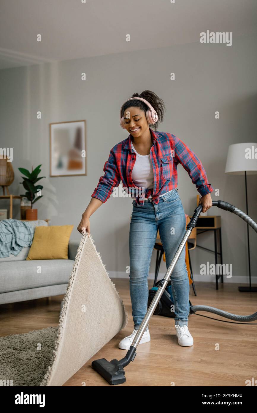 Cheerful young black female in wireless headphones with vacuum cleaner dusts floor, enjoys music in living room Stock Photo