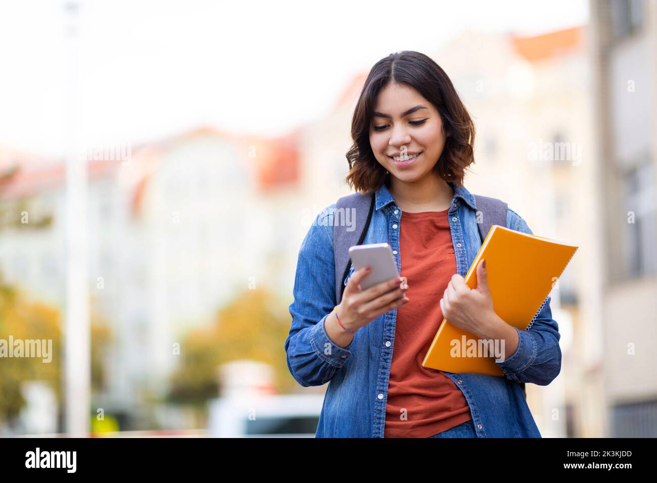 Middle Eastern Female Student Walking With Smartphone And Workbooks On City Street Stock Photo