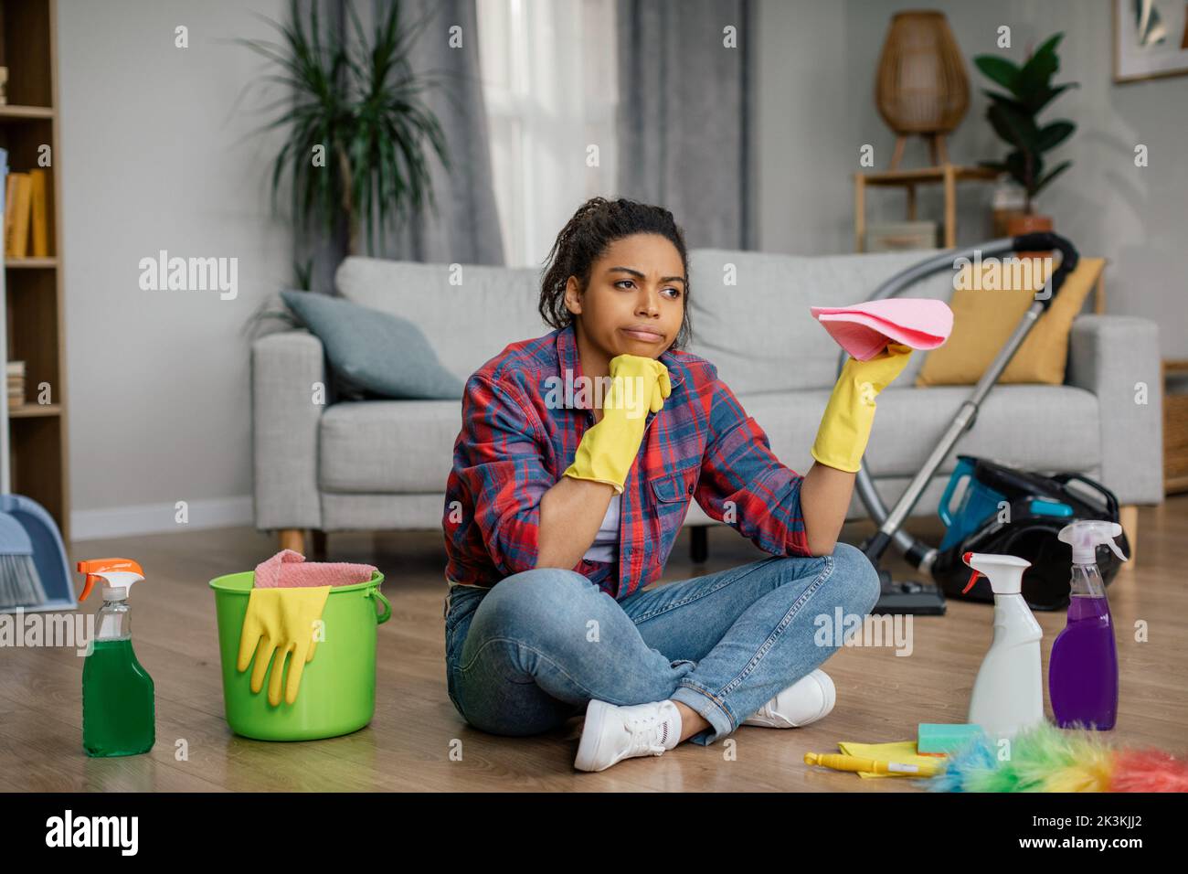 Sad bored young black woman in rubber gloves sits on floor with cleaning supplies, suffers from a lot of work Stock Photo