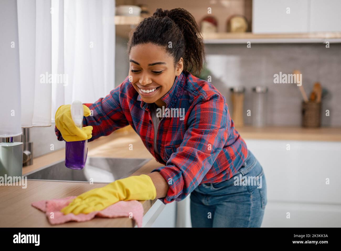 Cheerful young black woman wipes table from dirt with cleaning supplies in minimalist kitchen interior Stock Photo