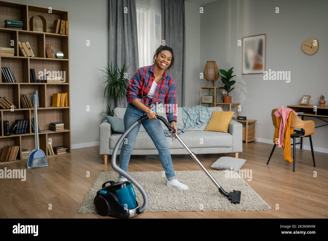 Smiling young black woman with vacuum cleaner dusts floor, enjoy cleaning alone in living room interior Stock Photo