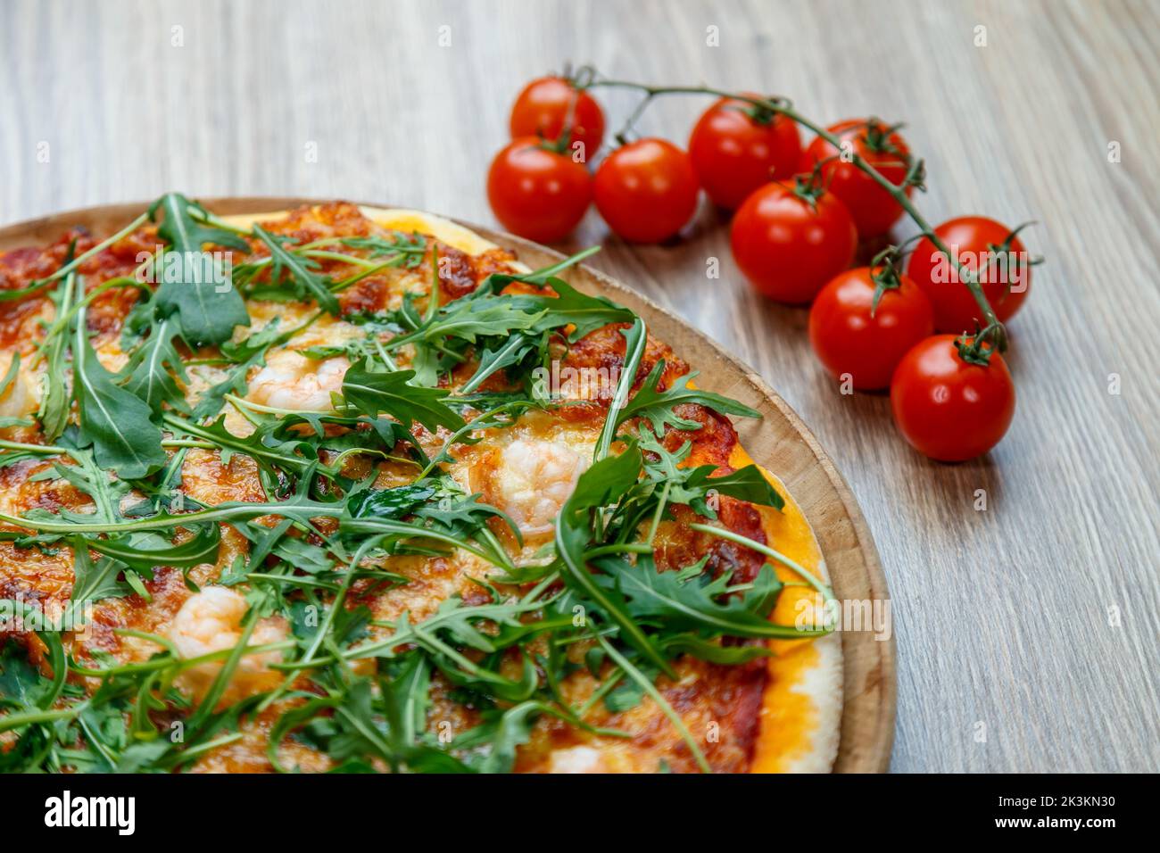 A closeup shot of a delicious gourmet pizza with arugula on a wooden table with a vine of tomatoes Stock Photo