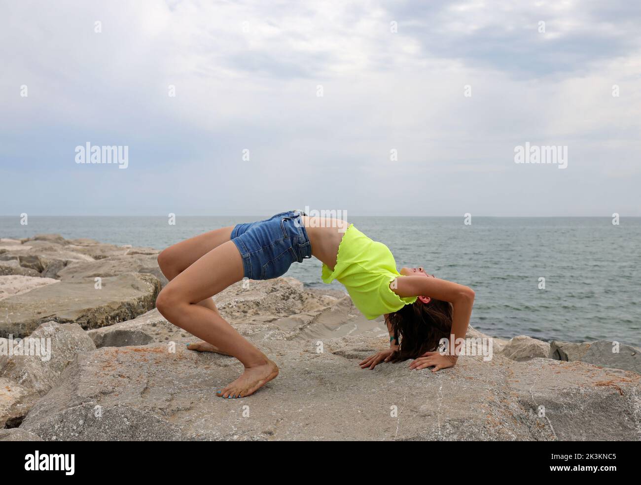 young girl performs gymnastic exercises arching her back backwards on the rocks by the sea without showing her face Stock Photo