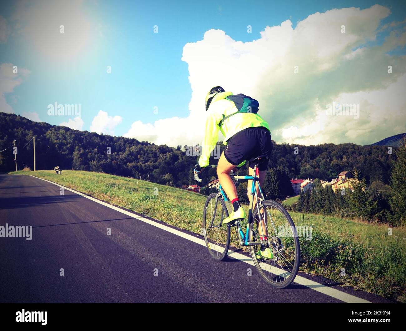 young cyclist with racing bicycle and waterproof jacket on mountain road rides fast seen from behind Stock Photo