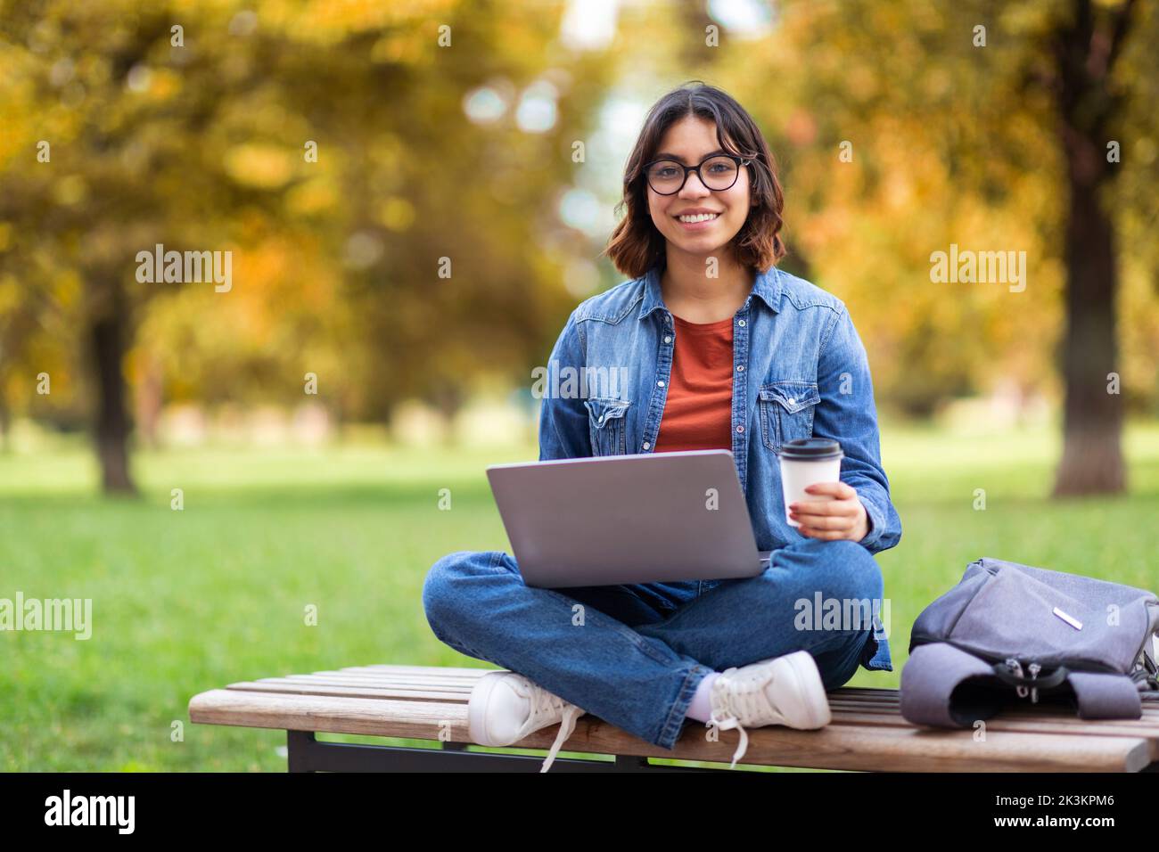 Smiling Young Middle Eastern Female Using Laptop And Drinking Coffee Outdoors Stock Photo