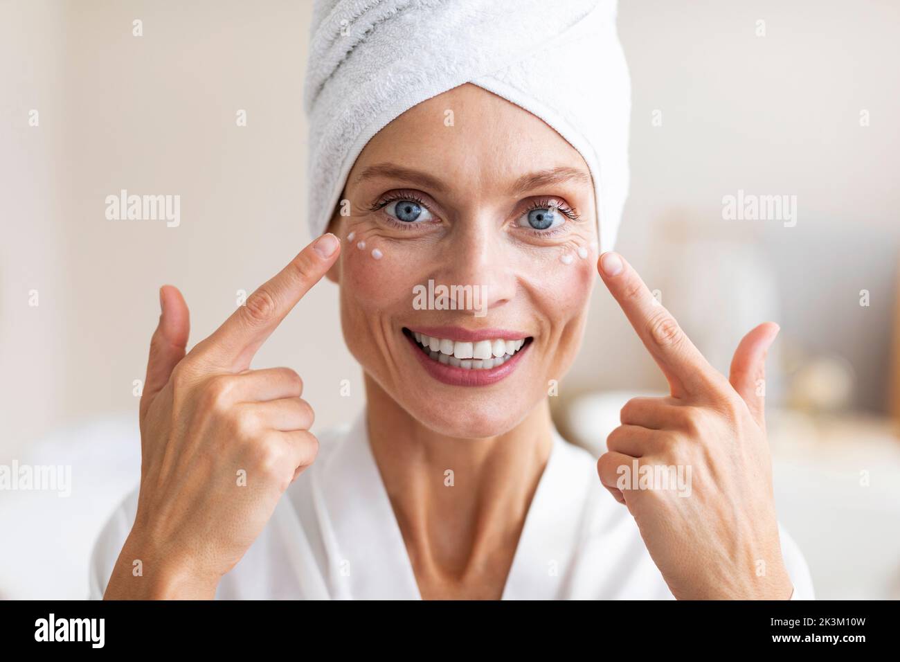 Mature beauty concept. Charming middle aged woman applying face cream under her eyes and smiling at camera Stock Photo
