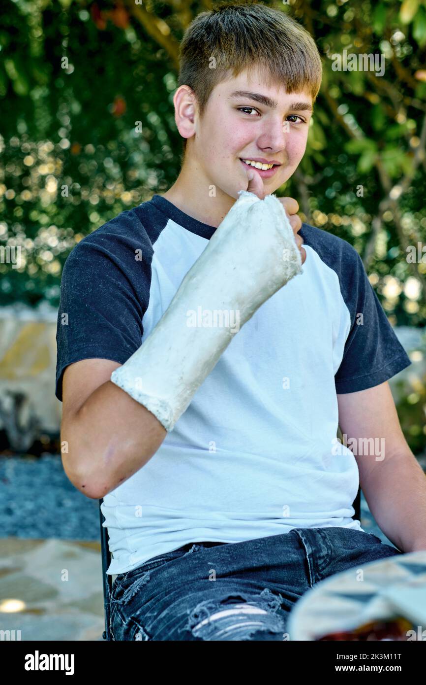 Portrait of young caucasian boy with a broken and cast arm sitting in a chair outdoor in a garden. Lifestyle concept. Stock Photo