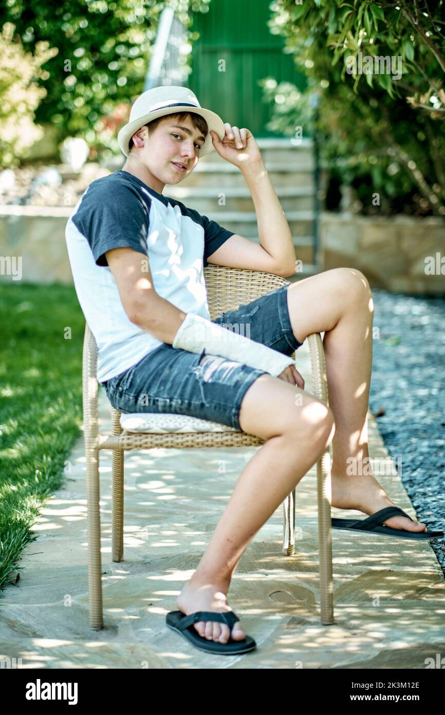 Portrait of young caucasian boy with a broken and cast arm wearing a hat and sitting in a chair outdoor in a garden. Lifestyle concept. Stock Photo