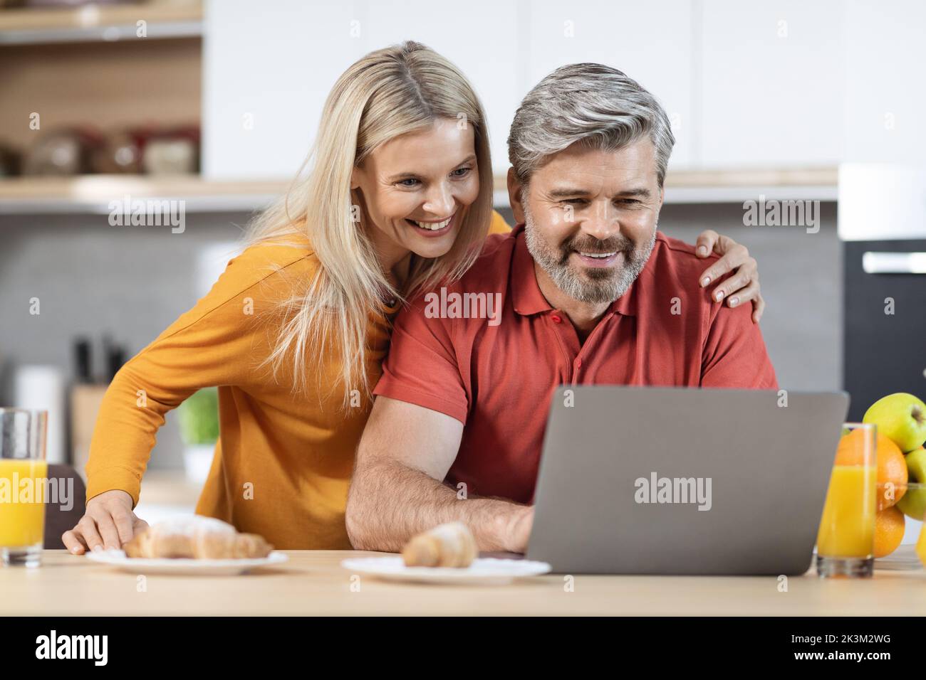 Happy spouses using laptop while having breakfast at kitchen Stock Photo