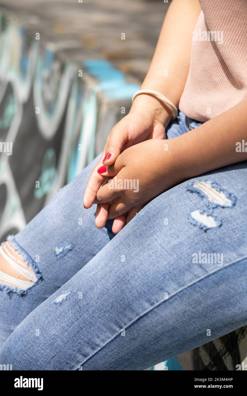 detail of the hands of a person with fingers crossed wears a bracelet and worn jeans, anonymous identity and nervous person gestures Stock Photo