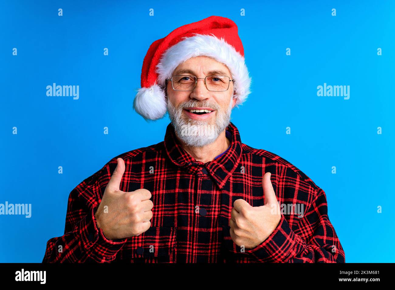 an elderly and well-groomed man in a plaid shirt and santa hat in blue studio background Stock Photo