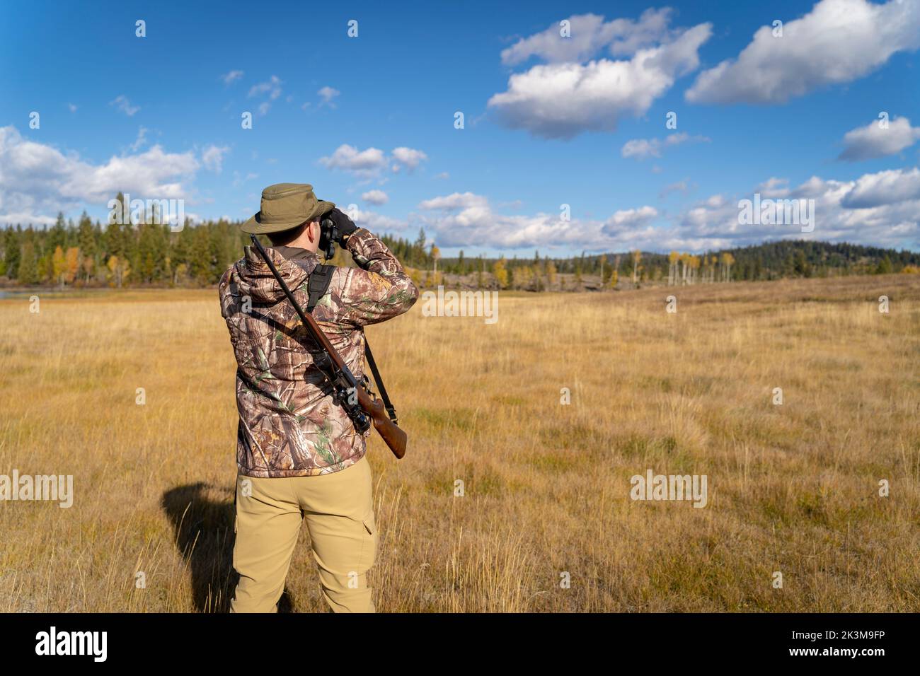 A sniper in camouflage and with a rifle looking through binoculars in a field Stock Photo