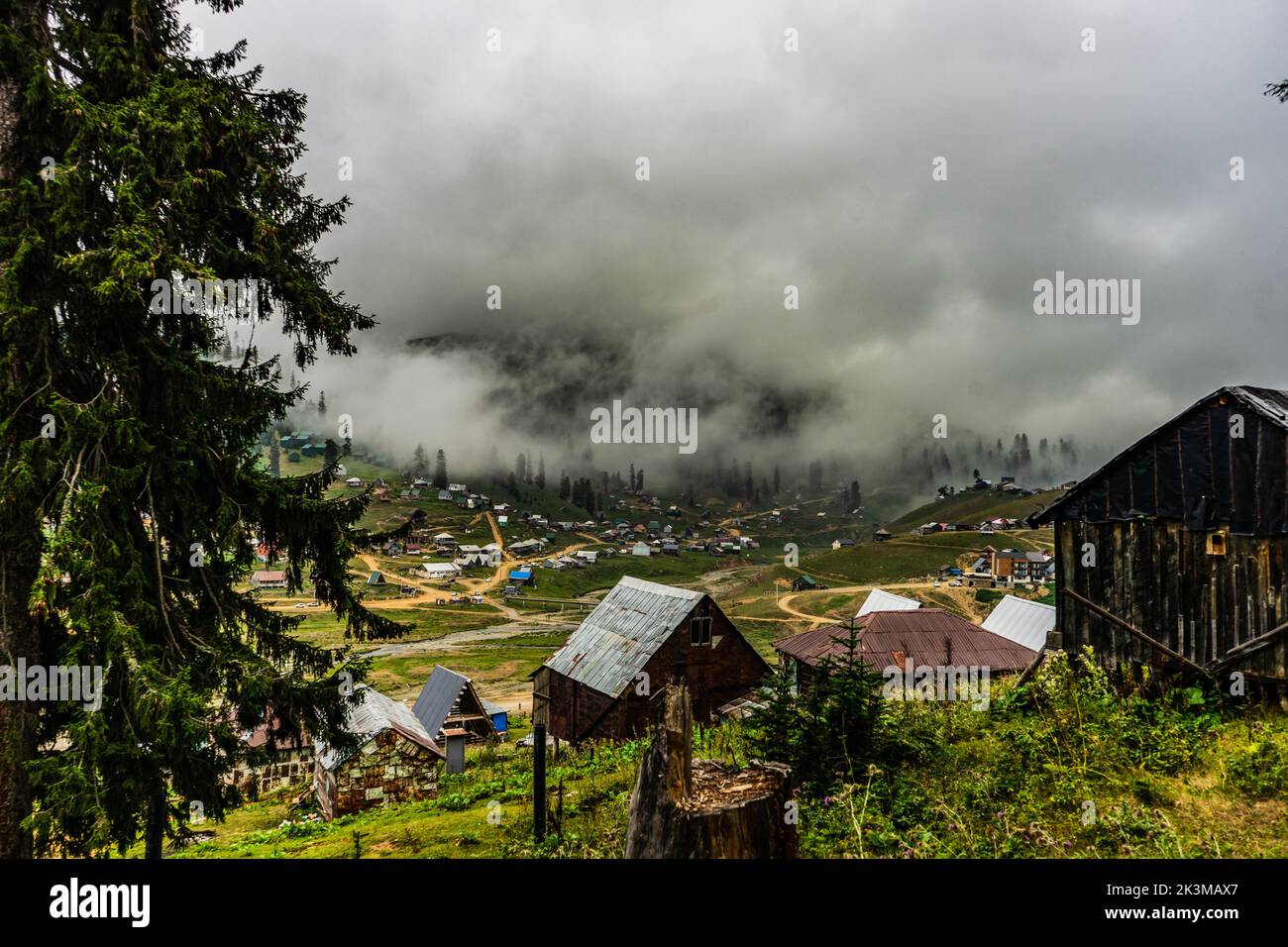 From above houses and coniferous trees located on green slope of mountain against blue sky with dark gray clouds and fog on stormy day in highlands of Stock Photo