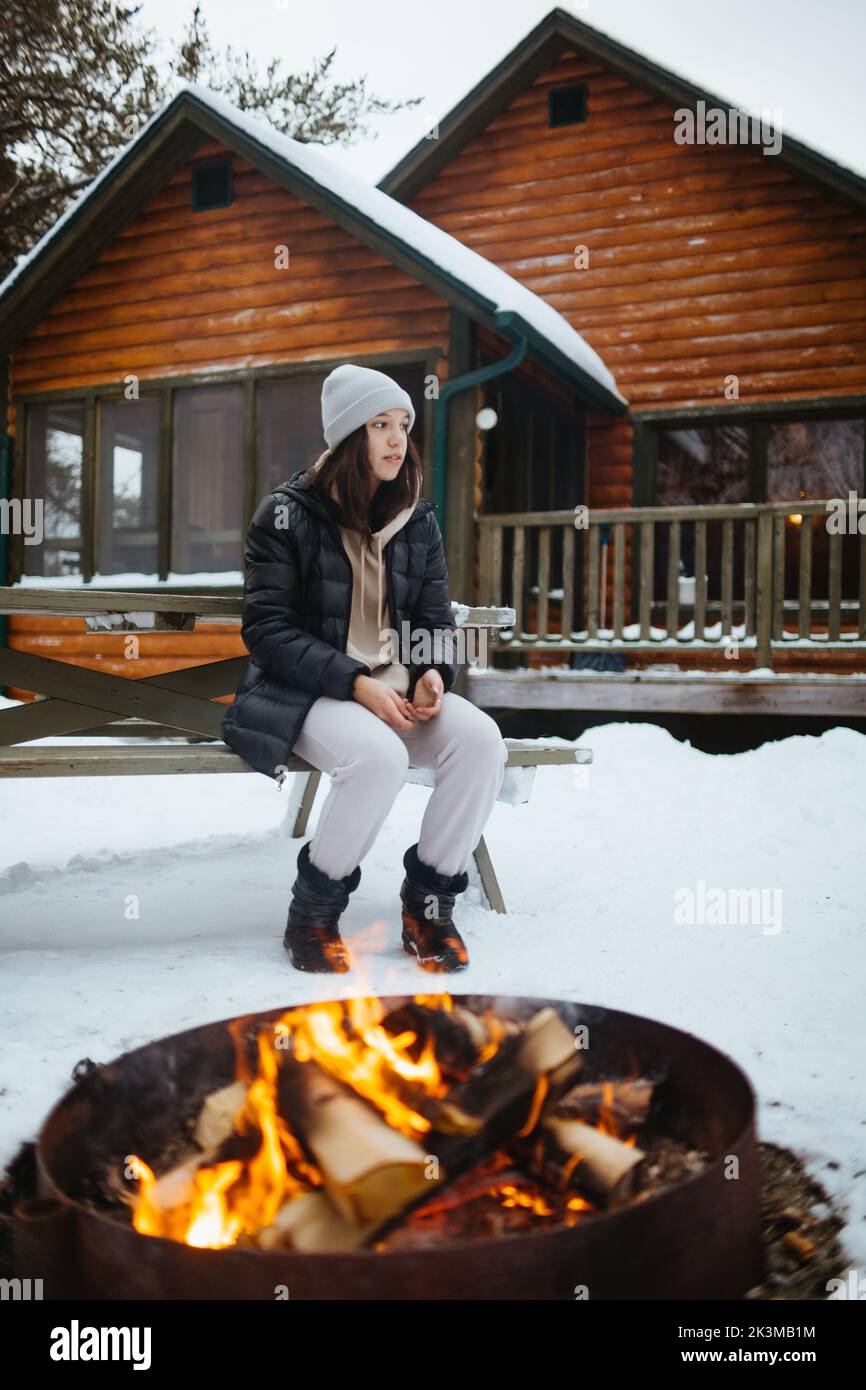 Full body woman in outerwear looking away while sitting on bench near brazier with burning logs outside wooden house on winter day in countryside Stock Photo