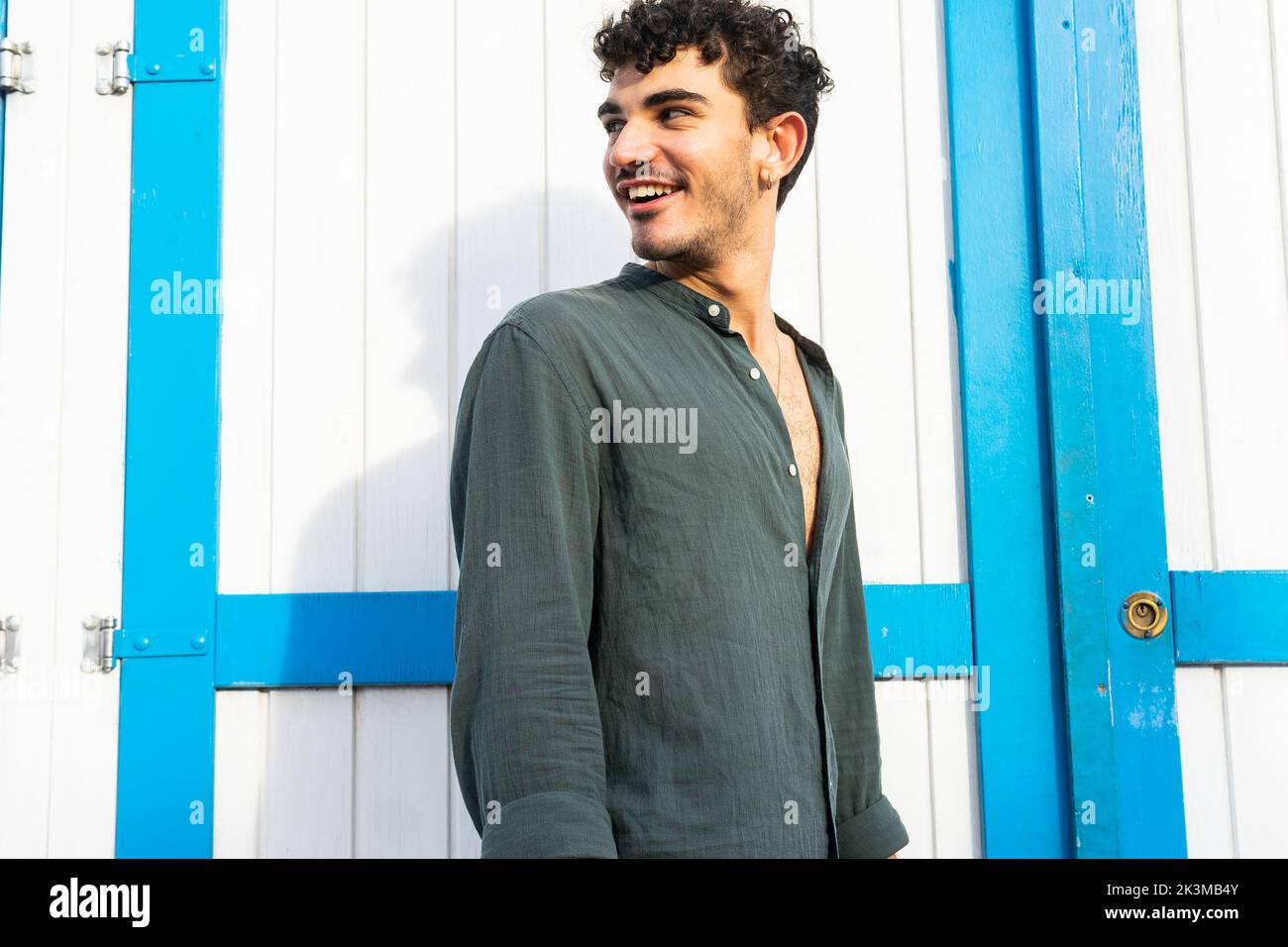 Young smiling Hispanic man in black shirt with curly hair looking away while standing near lumber door Stock Photo
