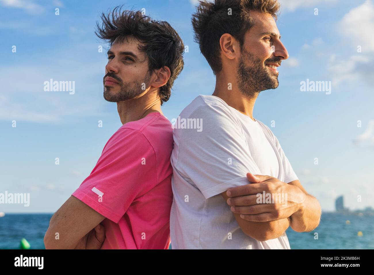 Confident Hispanic male athletes in t shirts crossing arms and looking away while standing back to back against sea and blue sky during break in fitne Stock Photo