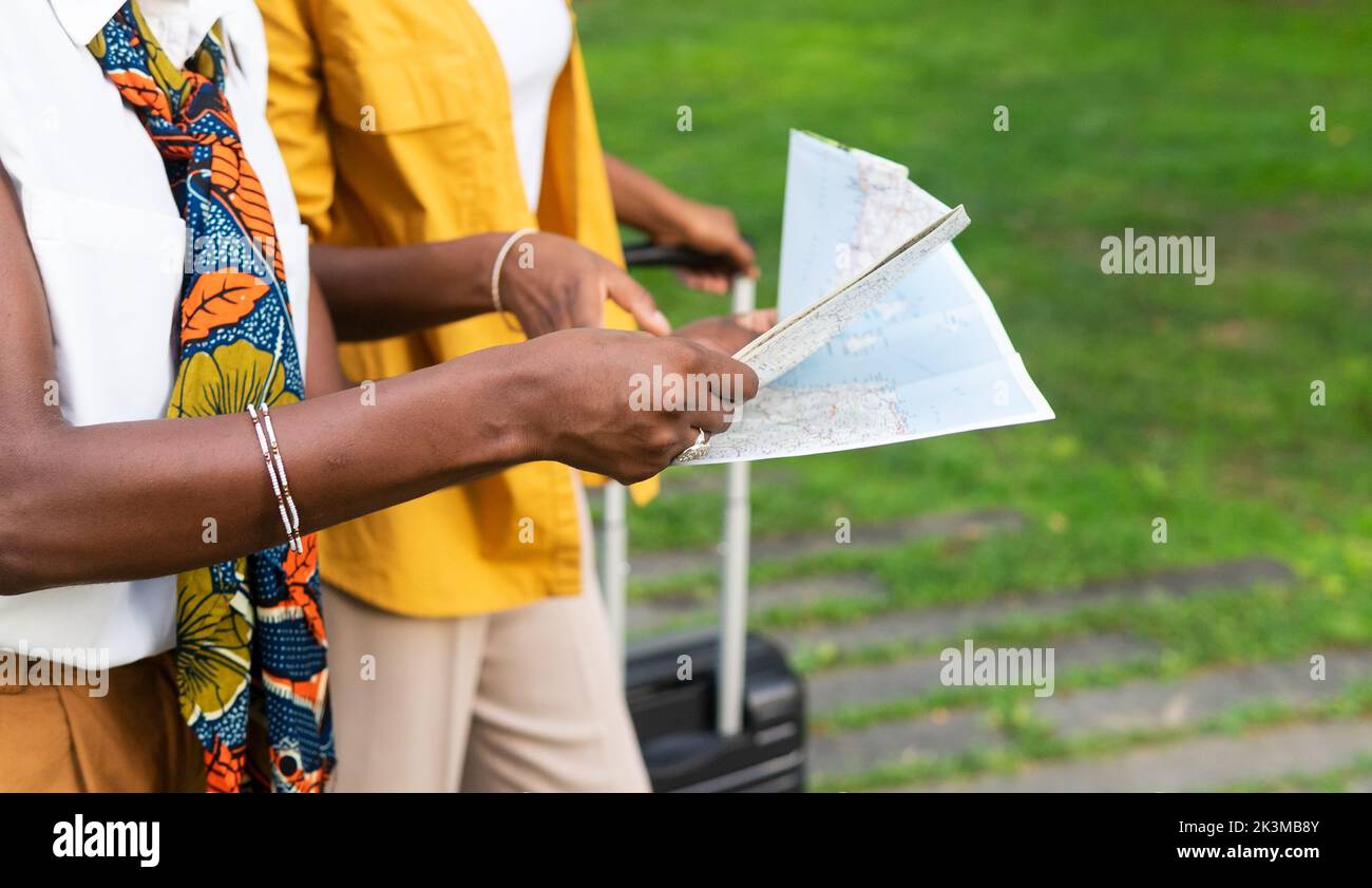 Crop young African American female friends in stylish clothes reading map while standing on street with suitcase during vacation Stock Photo