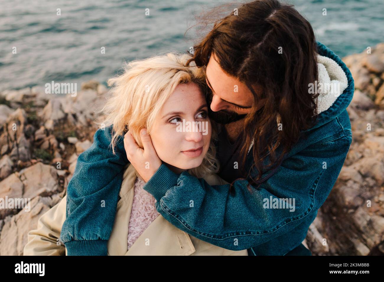 From above bearded man embracing blond girlfriend in outerwear and looking at each other while sitting on stony coast of rippling sea on cloudy day in Stock Photo