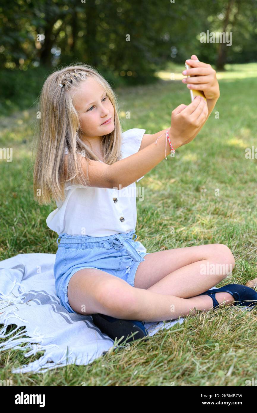 Full body blond girl in casual clothes sitting cross legged on blanket and taking selfie via smartphone while spending summer weekend day in meadow Stock Photo