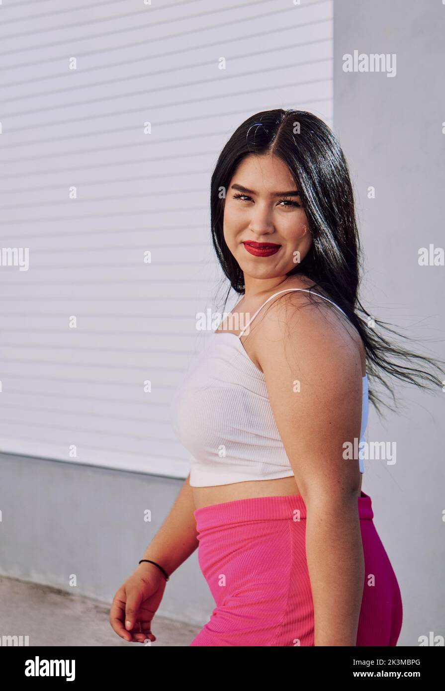 Side view of ethnic overweight female in stylish clothes with red lips smiling and looking at camera while walking against wall of modern building on Stock Photo