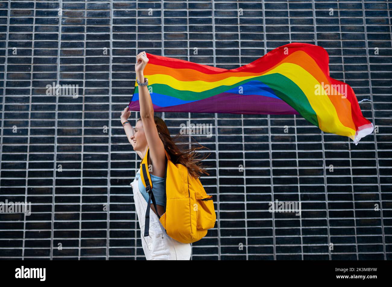 Low angle of young woman with backpack carrying rainbow flag in raised arms and running against black tiled wall during pride festival in city Stock Photo