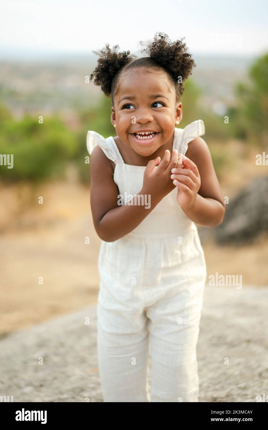 Optimistic little black girl in white clothes with hair buns looking away with smile on blurred