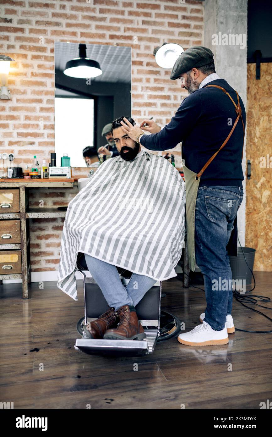 Ethnic hispanic customer in cape sitting on chair and getting haircut from professional barber working in grooming salon Stock Photo