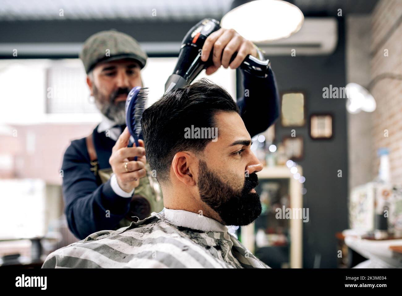 Concentrated mature ethnic barber styling dark hair of serious brutal client with dryer and comb in modern barbershop in daylight Stock Photo
