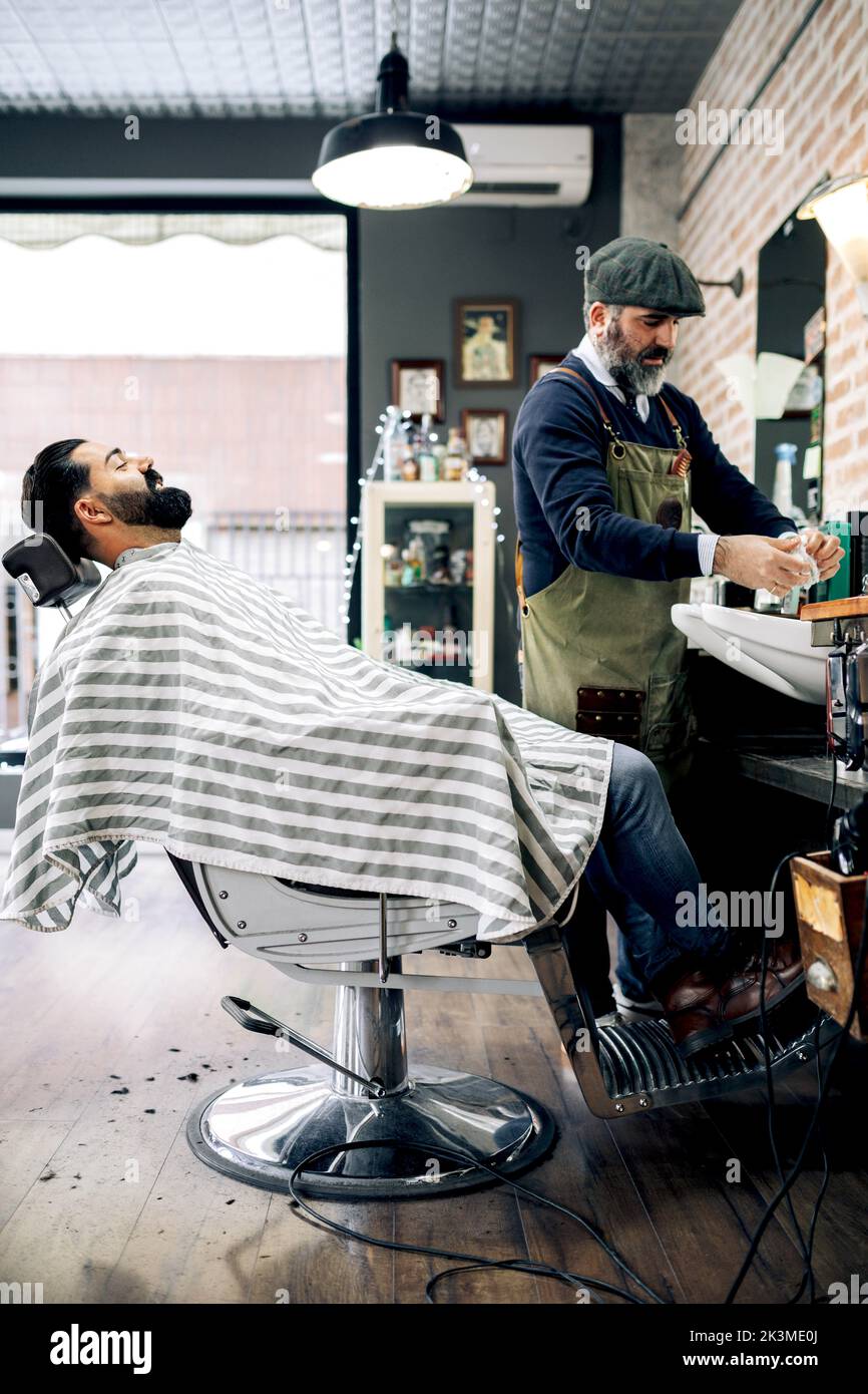 Side view of positive young bearded Hispanic male client sitting on chair near middle aged barber preparing for haircut in traditional barbershop Stock Photo