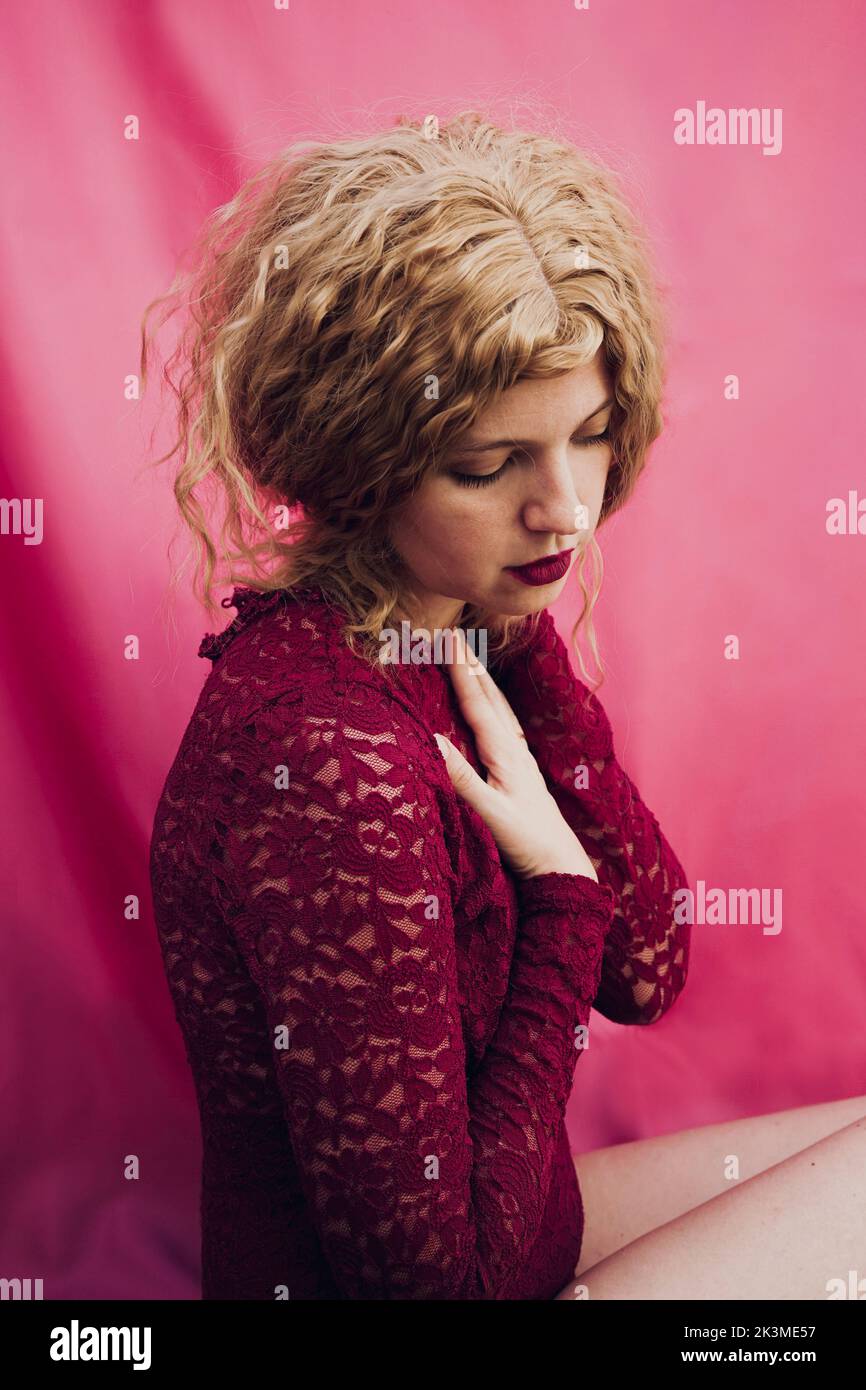 Side view of tranquil thoughtful female with bow in blond wavy hair sitting on pink textile background in studio Stock Photo