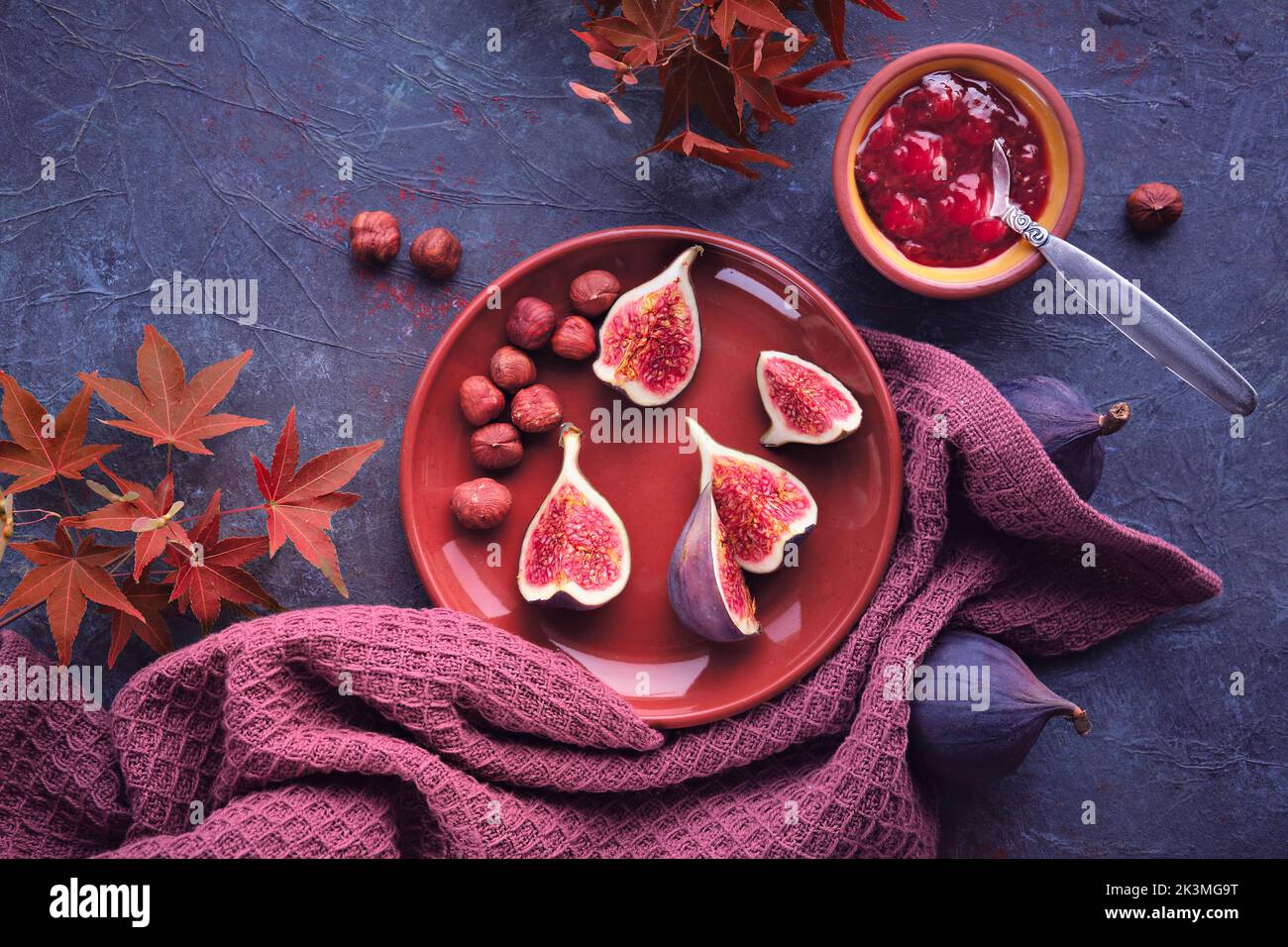 Autumntime background with fresh halved fig fruits on terracotta ceramic plate. Magenta towel and red maple Autumn leaves on dark purple board. Fig Stock Photo