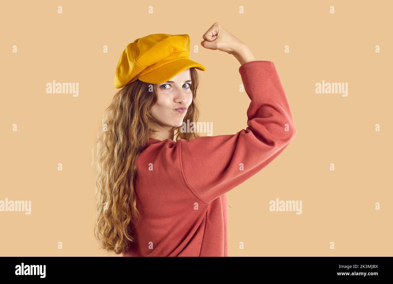 Confident young woman with wavy hair in yellow cap beret and red sweatshirt demonstrates power. Stock Photo